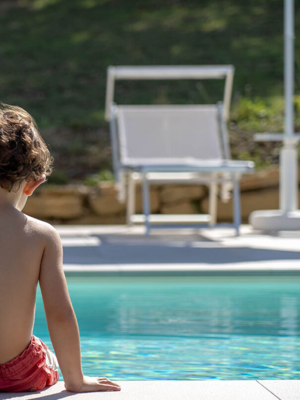 Bambino girato di spalle seduto a bordo piscina con le mani appoggiate sulle mattonelle del bordo. Di fronte a lui, la piscina e dall’altra parte della piscina, la zona solarium con ombrelloni e lettini