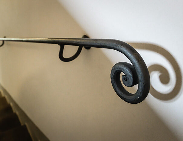 Dettaglio di un corrimano in ferro battuto che si trova sulla parete, lungo una rampa di scale. Nel dettaglio è presente un dettaglio del corrimano: un’arricciatura all’estremità.
