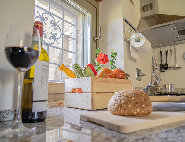 Foto ravvicinata che ritrae alcuni oggetti sopra il piano di una cucina. In primo piano sulla sinistra ci sono un valice di vino e una bottiglia di vino aperta; al centro in primo piano un tagliere con il pane. Al centro dell’immagine, tra la bottiglia di vino e il tagliere c’è una grande cassetta di legno con ortaggi (zucchine, pomodori e peperoni). Sullo sfondo della foto, si intravede il rubinetto del lavandino e una pentola appoggiata sui fornelli. Sulla parete accanto ai fornelli sono appesi dei mestoli da cucina e delle presine