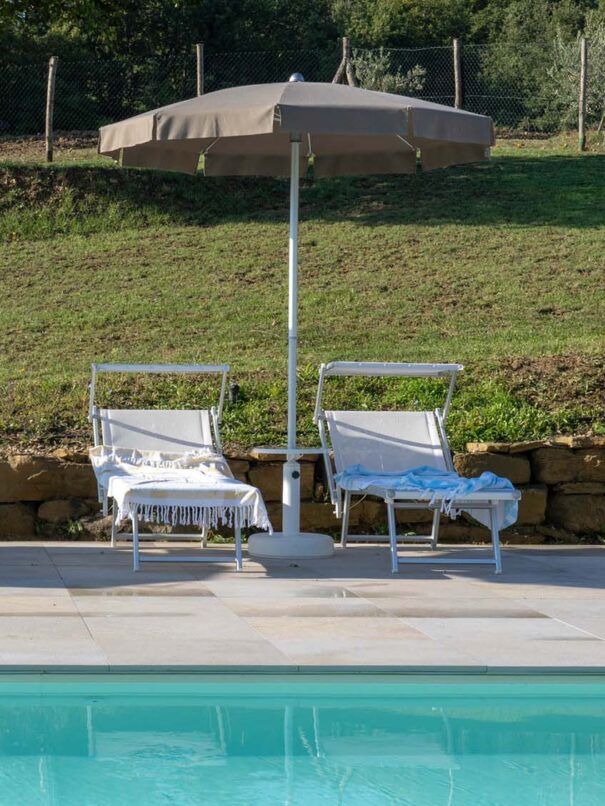Vista frontale dalla piscina sulla zona solarium che la circonda. Un pavimento con mattonelle circonda il bordo piscina e lì vi sono posizionati ombrelloni e sdraio in modo regolare e ordinato.