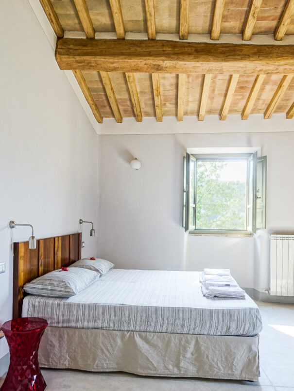 Foto dall’interno della stanza matrimoniale dell’Appartamento Le Ciliegie dell’Agriturismo Montelovesco. Sul lato sinistro dell’immagine, la testata in legno del letto matrimoniale poggia sulla parete della stanza e sopra di essa si trovano due luci da lettura appese al muro; accanto al letto si trova un comodino di forma rotonda. Sulla parete accanto al letto, al centro della foto, una finestra aperta illumina la stanza.