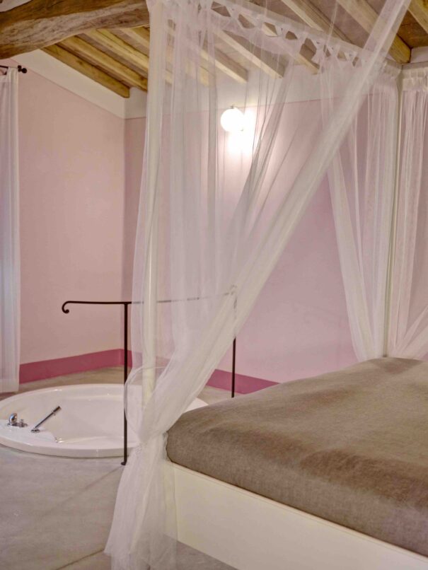 Al centro della foto, è posizionato un angolo del letto matrimoniale con tende che si trova all’interno della suite Le Lavande dell’Agriturismo Montelovesco. Ai piedi del letto, si trova una vasca da bagno a filo pavimento.