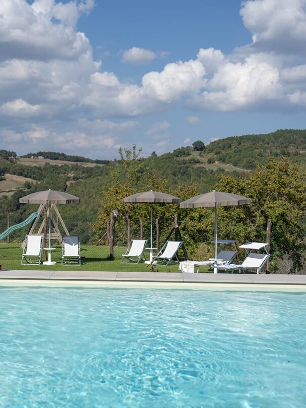 Vista della piscina dell'Agriturismo Montelovesco e del giardino in cui ci sono ombrelloni, sdraio e lettini.