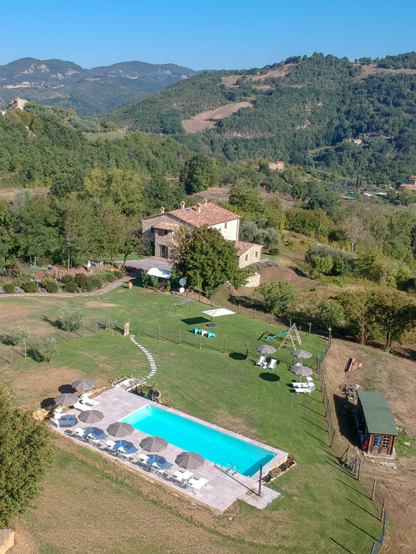 Foto panoramica dall’alto della piscina con del solarium dell’Agriturismo Montelovesco e dell'edificio. La piscina si trova all'interno di un grande giardino recintato. Nel lato superiore della foto c'è l'Agriturismo Montelovesco immerso nella natura.