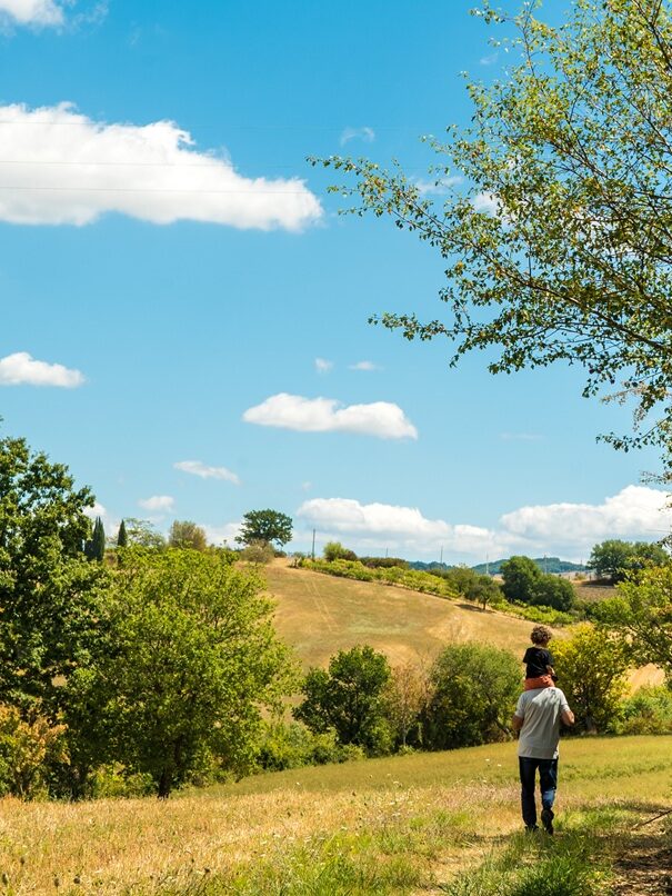 Foto panoramica del paesaggio circostante l'Agriturismo Montelovesco. Sul lato destro della foto si vede un uomo visto da dietro con un bambino sulle spalle, che cammina lungo i campi dell'Agriturismo Montelovesco. Sul lato sinistro si vedono terreni agricoli e colline.